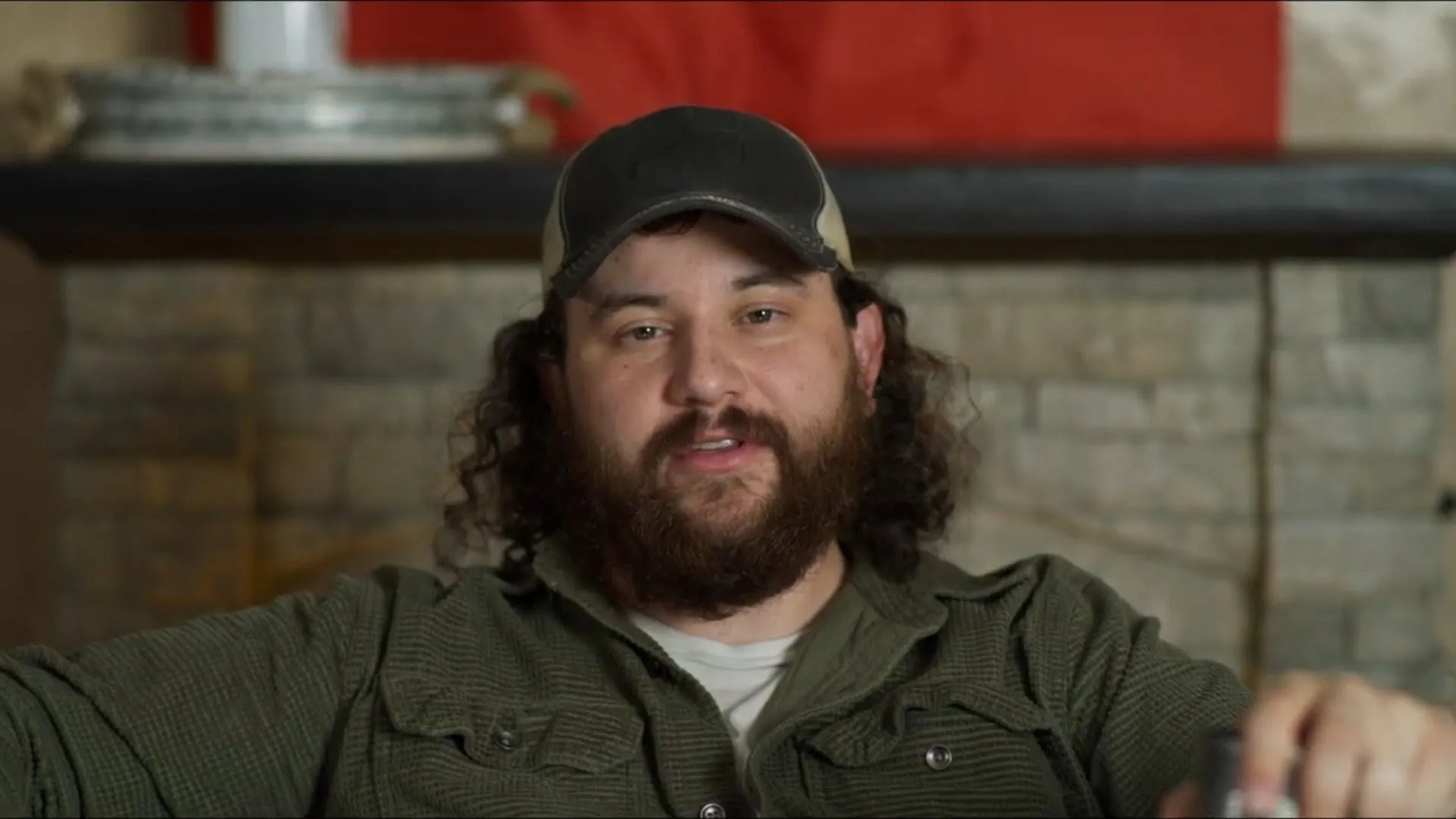 Tight interview frame of a bearded man in a cap and olive shirt speaking with a stone fireplace behind him